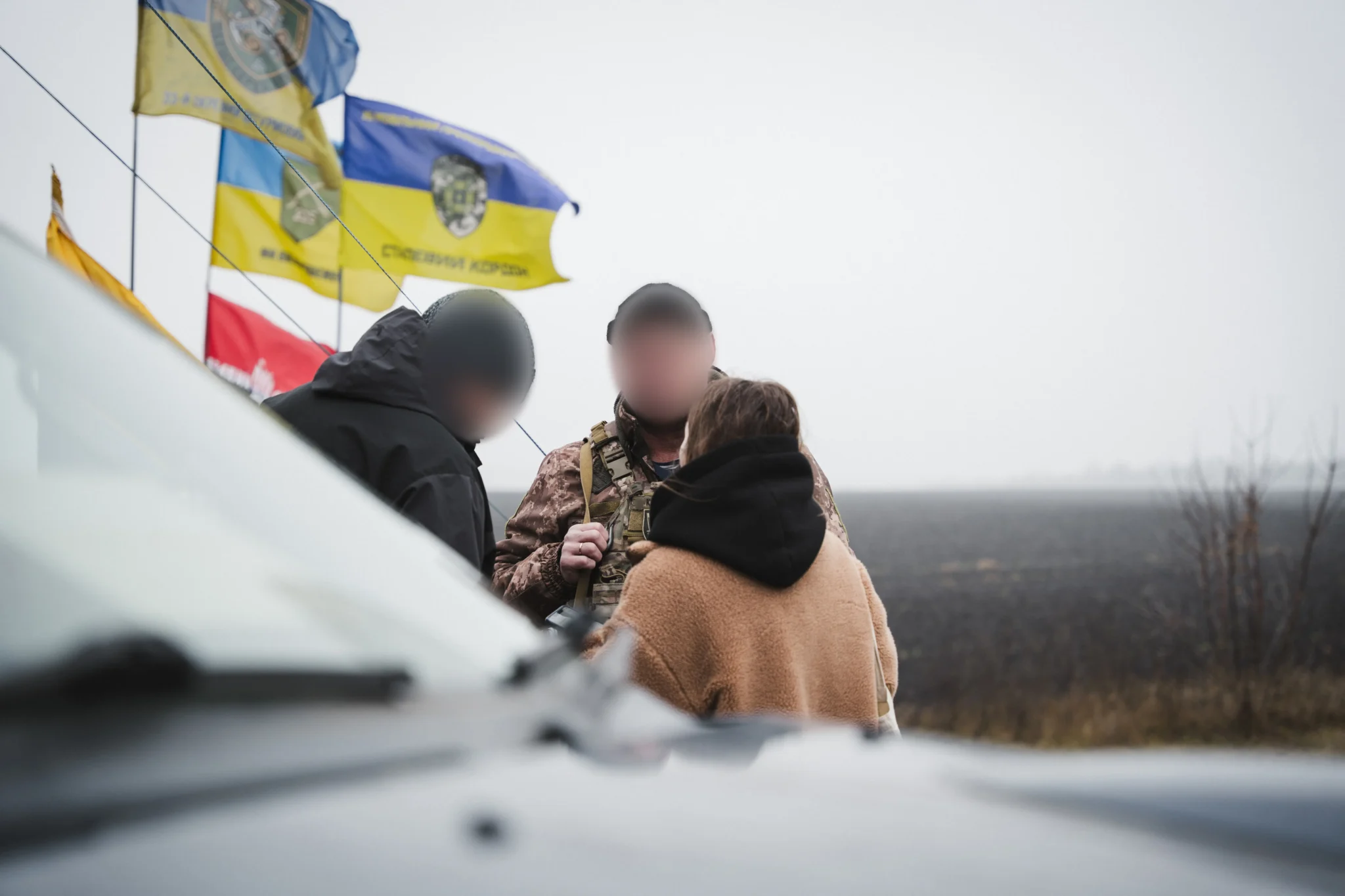 Журналістка спілкується з військовими поруч зі стелою. За нею — просторе поле, куди ступати небезпечно.