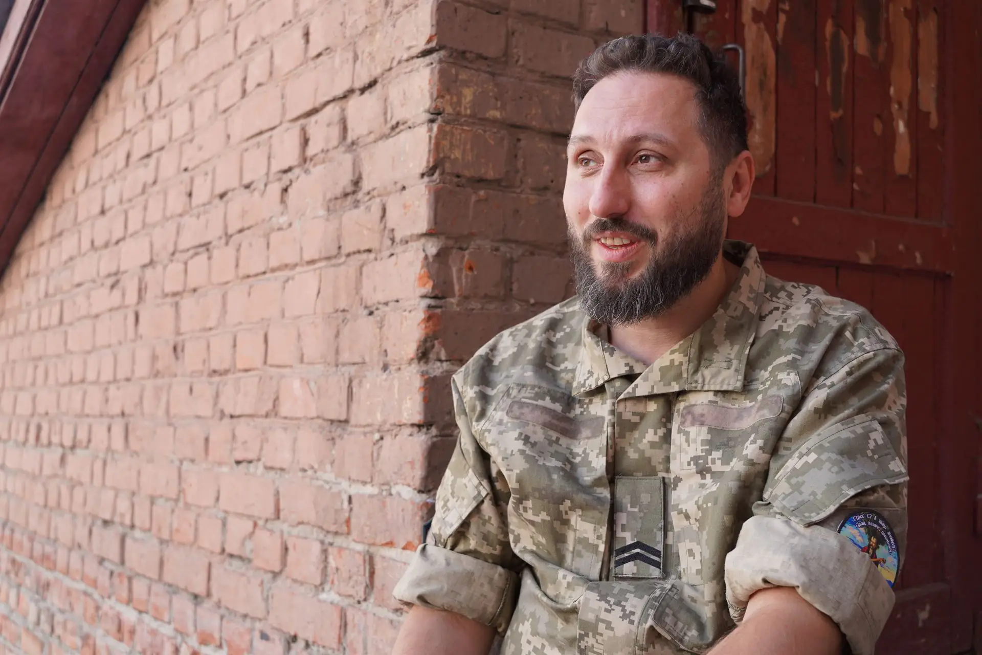 Військовослужбовець Роман Кандибур у формі сидить поряд з цегляним будинком. Він всміхається.